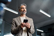 © insta_photos - Middle aged business man wearing suit using cellphone looking away in office. Mature older businessman executive leader wearing suit holding mobile phone at work standing with smartphone in hands.