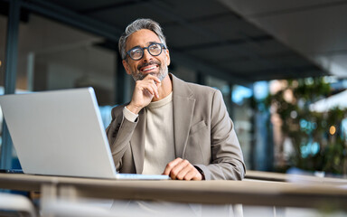 Wall Mural - Happy mature business man entrepreneur using laptop computer outside office looking away. Smiling busy middle aged professional male entrepreneur sitting at table outdoors hybrid working. Copy space