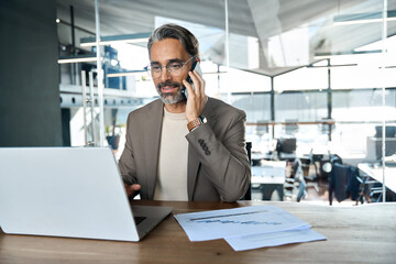 Wall Mural - Middle aged business man ceo talking on cell phone working on laptop in office. Busy mature professional businessman executive making call on cell using computer sitting at desk. Copy space.