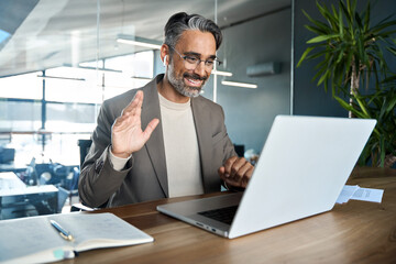 Wall Mural - Happy middle aged business man manager wearing earbud having virtual meeting looking at laptop hybrid working in office. Busy mature professional executive making video conference call greeting team.