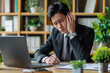 Overworked Japanese man having acute headache while working in office due to frustration, fatigue and crisis.