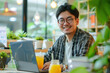Funny happy smiling Japanese man sitting at the desk on workplace at office with juice and booking tickets for summer vacation online via laptop.