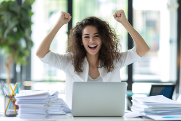 Wall Mural - Happy excited young Hispanic business woman accountant standing at the desk working on laptop computer with a pile of documents on table in office and making yes gesture rejoicing in successful job.