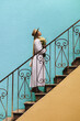 A girl in a white dress with flowers in her hands climbs the stairs against the background of a yellow-blue wall.