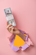 A woman's hand holds a stack of hundred dollar bills through a hole in a pink background.