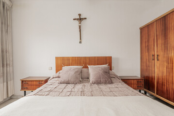 Wall Mural - bedroom with varnished wooden headboard and furniture, matching bedspreads and cushions and a Catholic cross on the wall