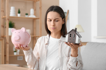 Canvas Print - Sceptical young woman with piggy bank and house model at home. Mortgage concept