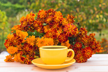 Wall Mural - Summer leisure activities concept; yellow cup of coffee and bunch of orange marigold flowers on a wooden table in green garden