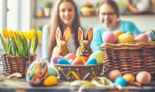 Two Smiling Women Watching Adorable Bunnies And Rattan Baskets Of Colored Chocolate Easter Eggs, Hand-painted Decoration, Cute Little Rabbits Next To A Basket Of April Yellow Tulips, Lovely Tradition