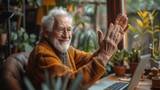 Fototapeta Big Ben - Elderly man with glasses smiles joyfully, waving at a laptop in a cozy room with plants