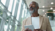 © Studio Nova - mature, professional man holding a digital tablet, looking contemplative, standing in a modern office environment with large windows.