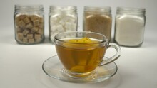 Brown Sugar Cubes Are Added In A Cup Of Tea Against The Background Of The Glass Jars With Various Types Of Sugar, Slow Motion