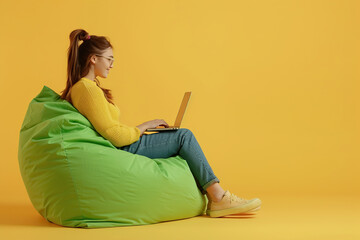 Attractive woman sitting on beanbag chair and using laptop on yellow background