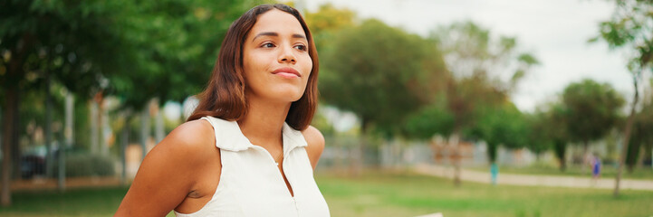 Wall Mural - Lovely tanned woman with long brown hair wearing white top sitting relaxed on park bench looking away, Panorama. Life style concept