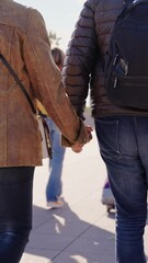 Wall Mural - Vertical unrecognizable adult tourist man and woman holding hands backwards while walking down the street with suitcase on vacation. Close-up of the bodies of two people from behind