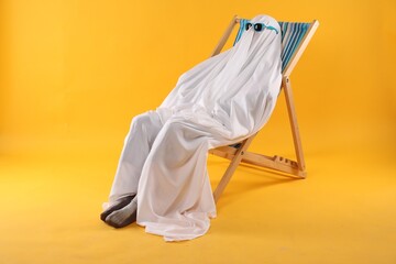 Person in ghost costume and sunglasses relaxing on deckchair against yellow background