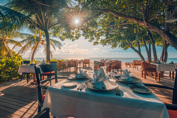 Wall Mural - Outdoor restaurant beach bar. Luxury table setting tropical beach restaurant. Sunset light trees wooden tables chairs under beautiful romantic vintage sky, sea view. Cozy hotel resort tourism travel