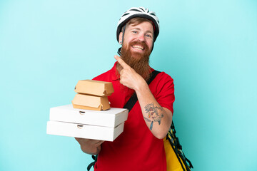 Wall Mural - Delivery man holding pizzas and burgers isolated on blue background pointing to the side to present a product