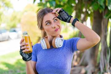 Wall Mural - Young pretty sport woman holding an orange juice at outdoors having doubts and with confuse face expression