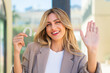 Pretty blonde Uruguayan woman holding invisible braces at outdoors saluting with hand with happy expression