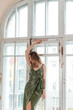 © bulashenko - A young stylish girl with white curly hair in a green dress stands on the windowsill of a large old window in the room