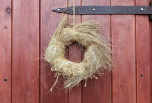 Wooden, Red Door With An Old Metal Hinge. A Round Wreath Of Dry Grass, Tied With Hemp Rope, Hangs On A Rusty Nail. Design.