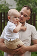 © Elena - A proud dad holds his happy one-year-old boy while sitting amidst lush green trees in the summer.