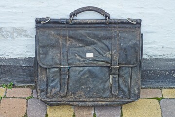 an old black big dirty tarpaulin  leather closed stylish  handbag with iron metal clasps stands  on a concrete surface near the white wall  in  the street 	
