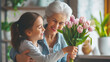 The granddaughter congratulates her beloved grandmother on spring holiday of March 8 or her birthday. Joyful senior woman receiving bouquet of pink tulip flowers from cute child
