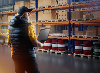 Wall Mural - Man in factory warehouse. Logistics center manager with laptop. Supervisor inspects warehouse racks. Man inside warehouse with barrels and boxes. Storage manager in cap and down vest