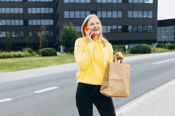 30s happy woman holding a paper bag in hands and calling on phone on city street. Technology, communication and people concept