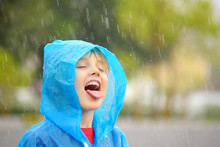 Portrait Of A Happy Child In The Pouring Rain. A Joyful Boy In A Blue Raincoat Catches Water Drops On His Tongue In The Pouring Rain While Walking.
