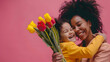 Little black daughter congratulating and hug mother and give her flowers for mothers day isolated on pink background. Happy ethnic family concept. Happy mother's day