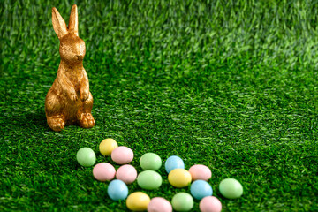 Wall Mural - Happy Easter, gold easter bunny and pastel colored candy coated Easter eggs on artificial green grass
