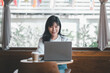 © Satori Studio - Business freelance concept, An engaging woman smiles warmly while working on a laptop in a bright and airy cafe, with a coffee cup at her side.