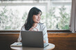 © Satori Studio - Business freelance concept, A hopeful woman engaging with her laptop in a spacious cafe, with the comfort of natural light from a large window.