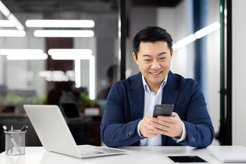 Wall Mural - Asian man inside office with phone in hands, smiling contentedly, businessman using smartphone sitting at desk, mature boss in business suit browsing social media, typing text message call.