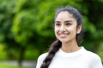 Wall Mural - Close-up photo of a happy young Indian woman standing outside the park, smiling, dreamy and contentedly looking to the side