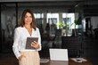 © Stock 4 You - Latin Hispanic mature adult professional business woman looking at camera and smiling. European businesswoman CEO holding digital tablet using fintech tab application standing at workplace in office.
