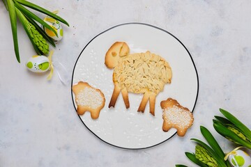 Easter pastries, a large lamb made of shortcrust pastry and two small lamb cookies on a white round ceramic board on a light concrete background with Easter decor.