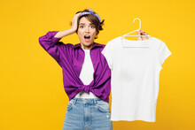 Young Shocked Scared Astonished Sad Woman Wears Purple Shirt Do Housework Tidy Up Hold T-shirt On Hanger Look Camera Put Hand On Head Isolated On Plain Yellow Background Studio. Housekeeping Concept.