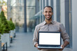 © Liubomir - Portrait of a smiling hispanic man holding a mockup of a laptop with a white screen, standing on the street near an office building and showing the device to the camera.