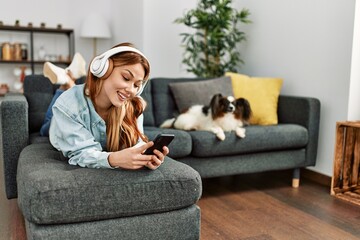 Wall Mural - Young caucasian woman listening to music lying on sofa with dog at home