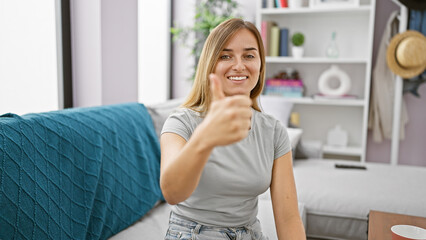 Sticker - Joyful, young blonde woman, sitting comfortably on the sofa, confidently flashes a positive thumb up gesture at home, radiating happiness in a cozy indoor setting.