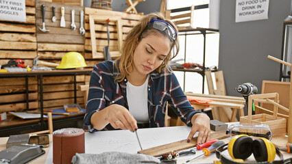 Sticker - Attractive young hispanic woman carpenter, dressed for safety with glasses, skillfully jots down notes in her carpentry workshop