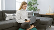 © Krakenimages.com - A blonde woman works on her laptop while seated comfortably in a modern living room, exuding casual elegance