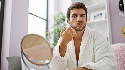 A young hispanic man in a white bathrobe grooming with a mirror at home, exhibiting casual morning routines and comfortable living.