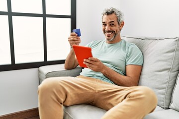 Wall Mural - Middle age grey-haired man using touchpad and credit card sitting on sofa at home