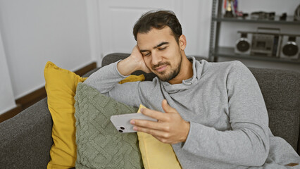 Wall Mural - Young hispanic man with beard using smartphone while relaxing on couch at home.