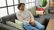 Relaxed young woman with dreadlocks sitting on modern gray sofa in a cozy, well-decorated living room